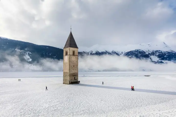 Torre sommersa nel lago di Resia, circondata da neve e montagne nebbiose. Alcune persone passeggiano sul ghiaccio.